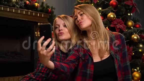 两个漂亮的年轻女孩或女人坐在新年或圣诞树附近用手机自拍视频的预览图