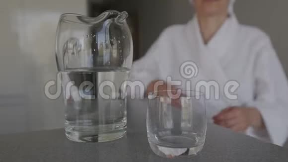 一个穿着白色长袍头上有一条毛巾的女人把水壶里的水倒进玻璃杯里喝视频的预览图