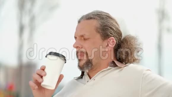一位放松迷人的留着长发的中年男子在绿色的长凳上用一次性杯子喝咖啡视频的预览图