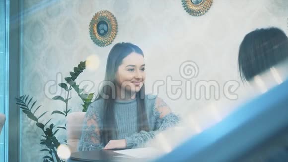 坐在咖啡馆里的漂亮年轻女人一个穿着休闲服装的白人女孩在窗户玻璃上的漂亮而温暖的镜头视频的预览图