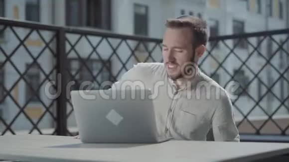肖像自信胡须的年轻人坐在笔记本电脑前的露台上的桌子旁工作概念视频的预览图