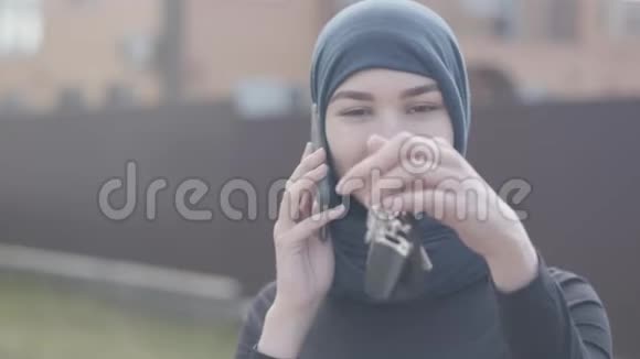 独立微笑的年轻穆斯林妇女戴着传统的头巾手里拿着手机和汽车钥匙的画像视频的预览图