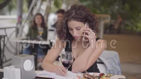 美丽的卷发女孩写下了坐在咖啡馆的桌子旁用手机聊天的信息忙碌的自由职业者视频的预览图