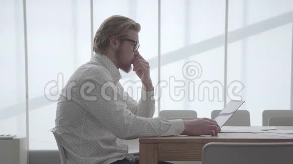金发体贴的人戴着眼镜坐在桌旁在上网本前的一间舒适的办公室里打字视频的预览图