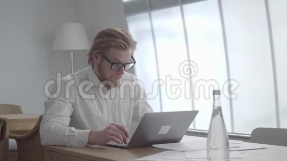 自信的金发体贴的男人戴着眼镜坐在上网本前一间舒适的办公室里的桌子旁视频的预览图