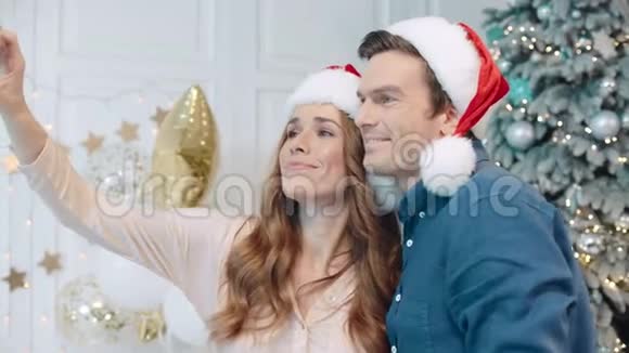 微笑的夫妇在圣诞树附近的圣诞老人帽子里自拍视频的预览图