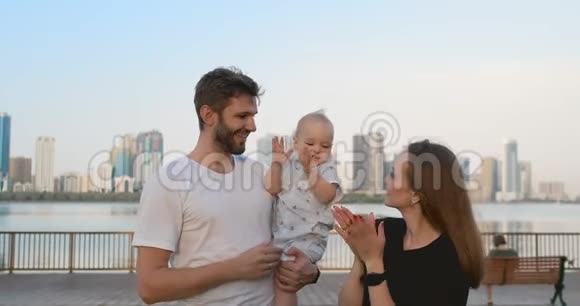 城市的父母爸爸妈妈抚养她的儿子站在城市的背景下的海滨视频的预览图