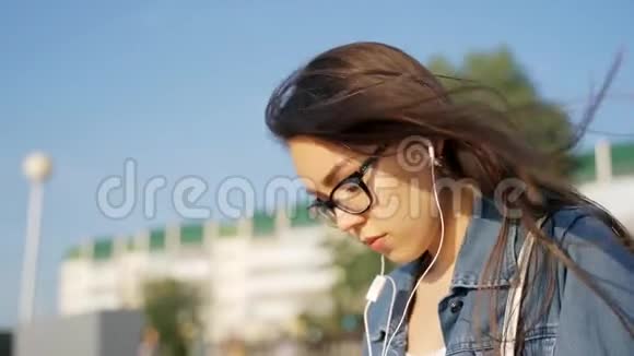 一个戴眼镜的年轻女孩坐在街上用耳机听音乐随风飘扬的头发特写视频的预览图