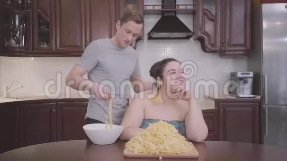 肖像可爱丰满的女人坐在家里的厨房里那个苗条的金发男人把面条挂在他肥胖的耳朵上视频的预览图