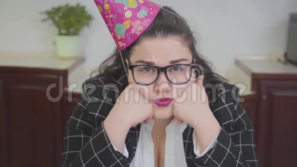 特写肖像悲伤丰满的女人戴着生日帽和时尚的眼镜坐在厨房的桌子旁胖子视频的预览图