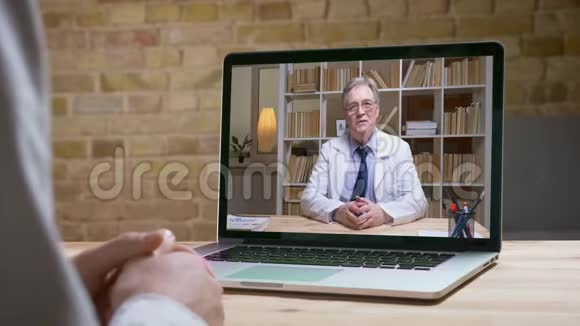 戴着白色实验室外套和眼镜的资深医生在网上视频卡上与屏幕后的病人认真交谈视频的预览图
