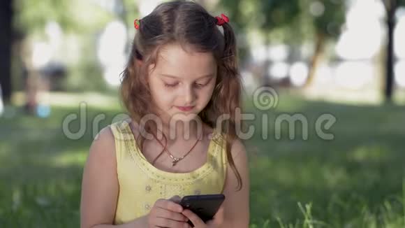 在阳光明媚的夏日一个女孩坐在公园的草地上用智能手机与社交网络中的朋友交流微笑视频的预览图