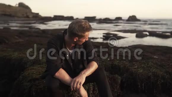 英俊的年轻人穿着时尚的深色衣服坐在黑色的海滩上享受着日落视频的预览图
