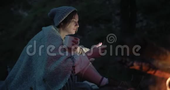 黄昏时一个年轻的女人坐在火对面的森林里用智能手机视频的预览图