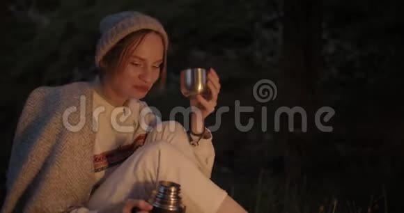 一个年轻的女人晚上坐在火对面的森林里从热水瓶里倒茶喝视频的预览图