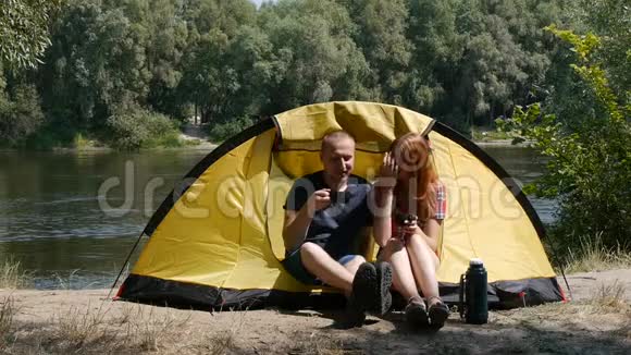 年轻的幸福夫妇坐在帐篷里看风景喝茶笑着说话背景上的森林和河流视频的预览图