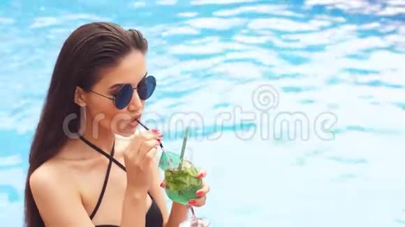 穿着时髦泳衣和太阳镜的魅力女孩躺在泳池边喝鸡尾酒视频的预览图