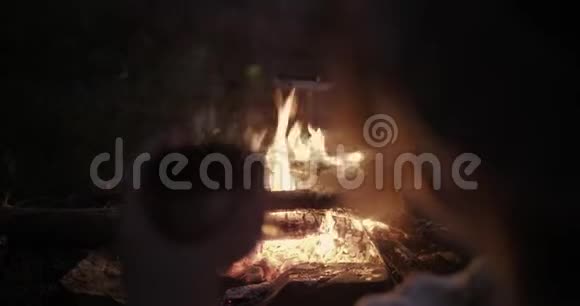 一个年轻的女人晚上坐在篝火前喝着茶看着篝火视频的预览图
