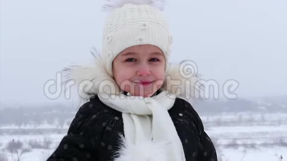 特写一个戴着白色针织帽子的小女孩看着镜头美丽地微笑着街上正在下雪她看起来视频的预览图