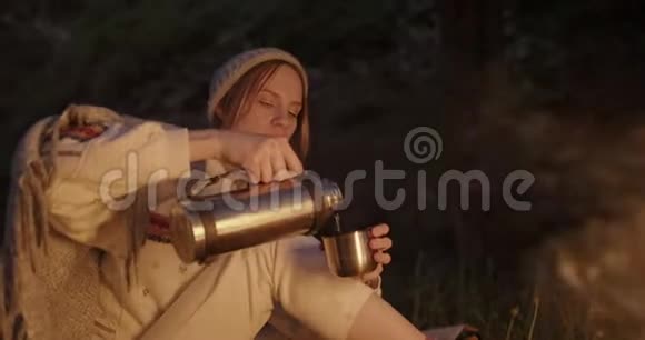 一个年轻的女人晚上坐在火对面的森林里从热水瓶里倒茶喝视频的预览图