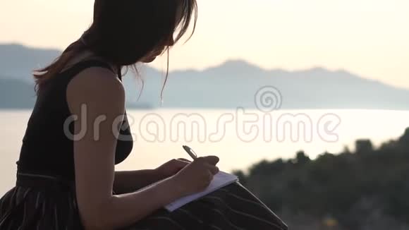 年轻女子在笔记本上写字日出时坐在海边慢动作视频的预览图