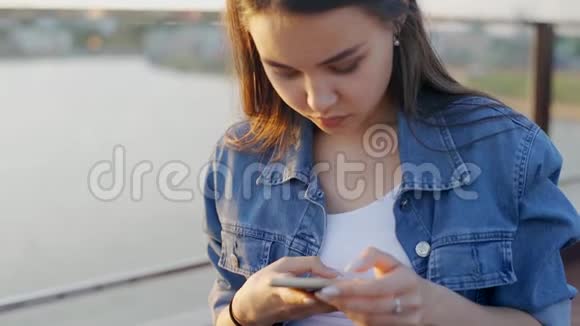 穿牛仔夹克的女孩正在智能手机上键入一条短信那个拿着电话的女孩在咖啡馆里户外视频的预览图