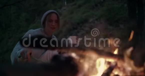 黄昏时分一个年轻的女人坐在火对面的森林里用木棍玩煤视频的预览图