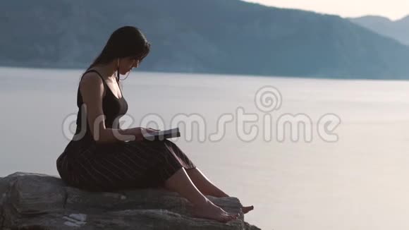 美丽的女孩读一本书赤脚坐在靠近大海的岩石上慢动作视频的预览图