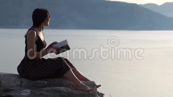 浪漫的女孩光着脚坐在海边和山上的岩石上视频的预览图