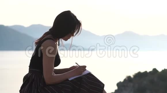美丽的女人在笔记本上写字日出时坐在海边慢动作视频的预览图