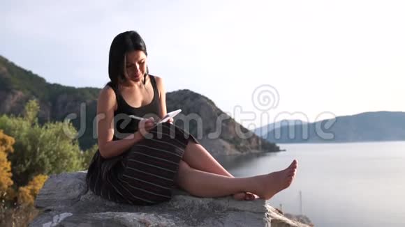 一个性感的女孩穿着笔记本光着脚坐在海面上的岩石上视频的预览图