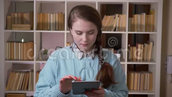 一位年轻漂亮的女学生在平板电脑上发短信看着大学图书馆里的照相机特写照片视频的预览图