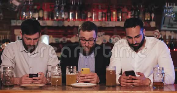 三个老朋友坐在啤酒酒吧里用智能手机有胡子的男人在他的智能手机上展示有趣的东西视频的预览图