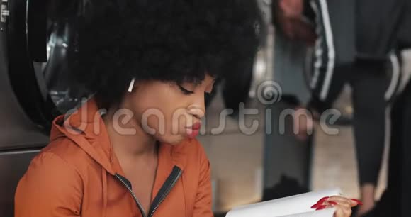 漂亮的非洲裔美国年轻女性在洗衣店洗衣服时看书她在等待视频的预览图