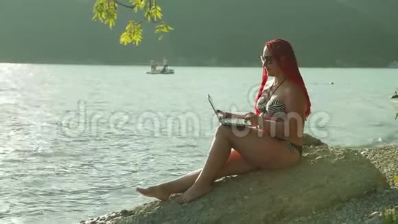 一个红头发的女人在岸边贴着群山的背景写博客视频的预览图