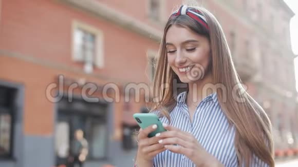微笑的年轻女子穿着蓝色和白色条纹连衣裙使用智能手机站在老街的背景上视频的预览图