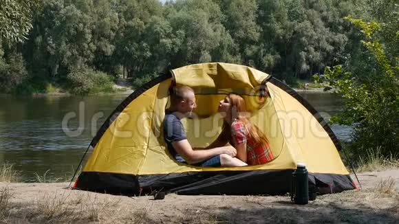在帐篷里的一对幸福的年轻夫妇女孩吻一个男人他们有一个浪漫的时刻森林和河流的背景视频的预览图