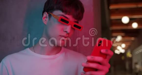 时髦的现代派眼镜中年轻的MillenialGuy站在一边躺在墙上用他的智能手机期待着视频的预览图