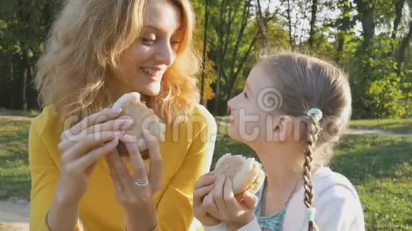 快乐的年轻金发母亲和她的小女儿正在公园里坐在长凳上吃快餐视频的预览图