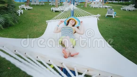 一个穿着酷色衣服和一顶宽边帽子的聪明女孩正坐在白色纺织吊床上的海边视频的预览图