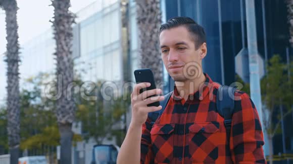 穿着格子衬衫背着背包在手机上聊天浏览着现代人身边的英俊白种人视频的预览图