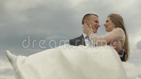 新郎把新娘抱在天空背景上新婚夫妇幸福家庭视频的预览图