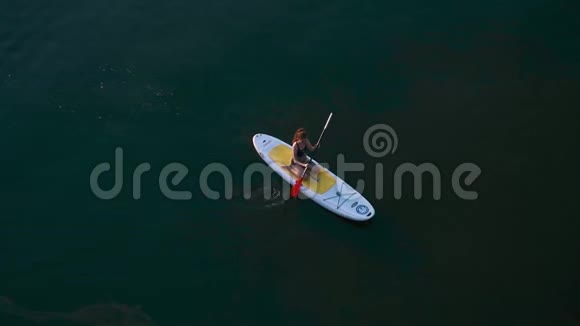 鸟瞰妇女坐在一个冲浪板上划桨划艇在一条光滑平静的河流晚上在河上水视频的预览图