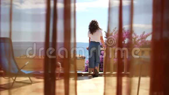 海边一个阳光明媚的露台上的女孩向远处的人挥手一只狗在附近散步从后面看视频的预览图