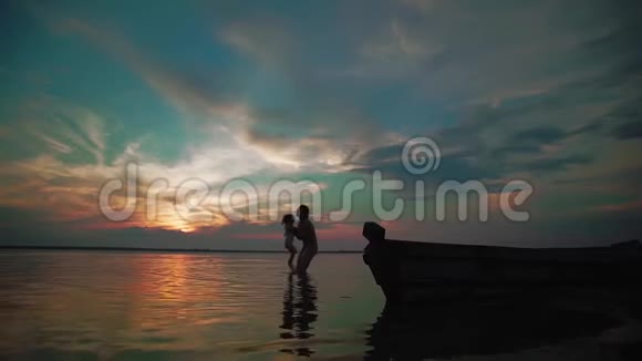 慢动作一个女人把孩子从湖边抱起来在日落时和她一起在湖边快乐地转过身剪影式视频的预览图