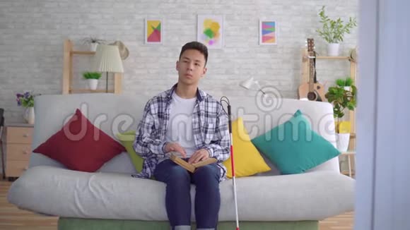 盲人亚洲年轻人坐在客厅的沙发上看一本盲文书视频的预览图