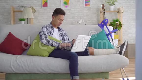 戴着绷带的断腿亚洲年轻人躺在沙发上手提电脑视频的预览图