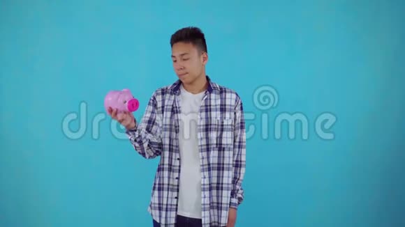 一个困惑的亚洲年轻人拿着蓝色背景的储蓄罐视频的预览图
