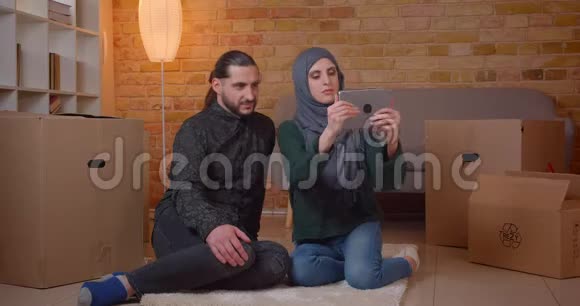 一对年轻快乐的穆斯林夫妇在一间新买的公寓里坐在盒子旁边的地板上用这张照片拍摄特写镜头视频的预览图