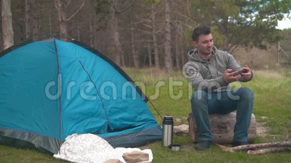 一个年轻人坐在蓝色帐篷附近的一根大木头上在电话里自拍视频的预览图